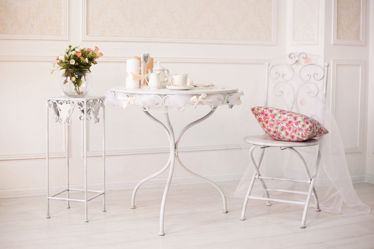 A white room with flowers on a table, a tea set on a table, and a flower print pillow on a chair