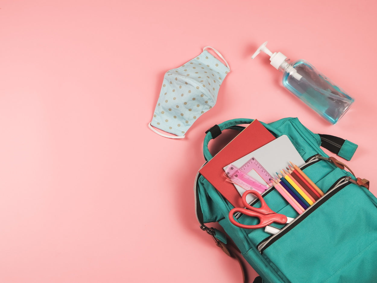 Backpack filled with school supplies and a polka dot facemask against a pink background