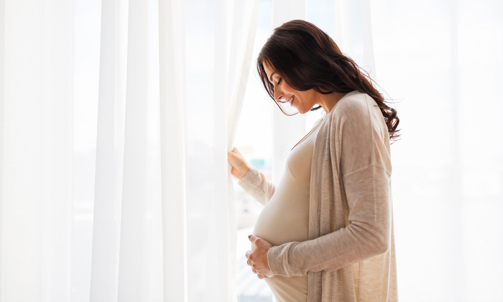 A pregnant woman stands by a bright window with curtains