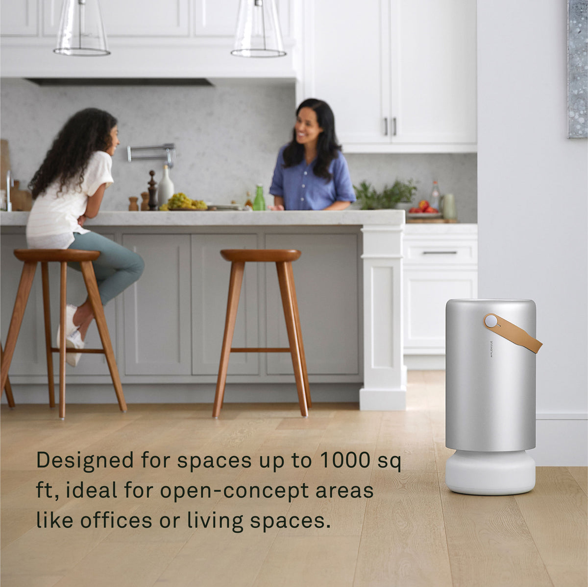 A mother and daughter talk at a kitchen island in their modern home. A Molekule Air Pro air purifier is nearby. Image text reads: Designed for spaces up to 1000 sq ft, ideal for open-concept areas like offices or living spaces.