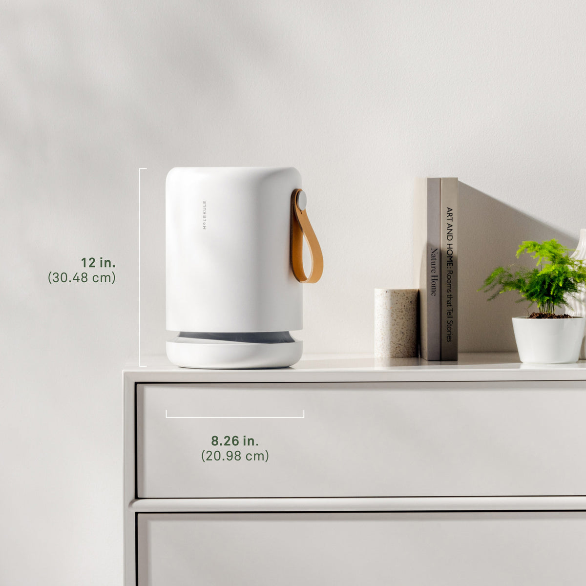 A Molekule Air Mini+ air purifier sits on a modern white credenza, coordinating with a stack of books and a small succulent. It stands 12 inches (30.48cm) tall and 8.26 inches (20.98cm) wide and deep.